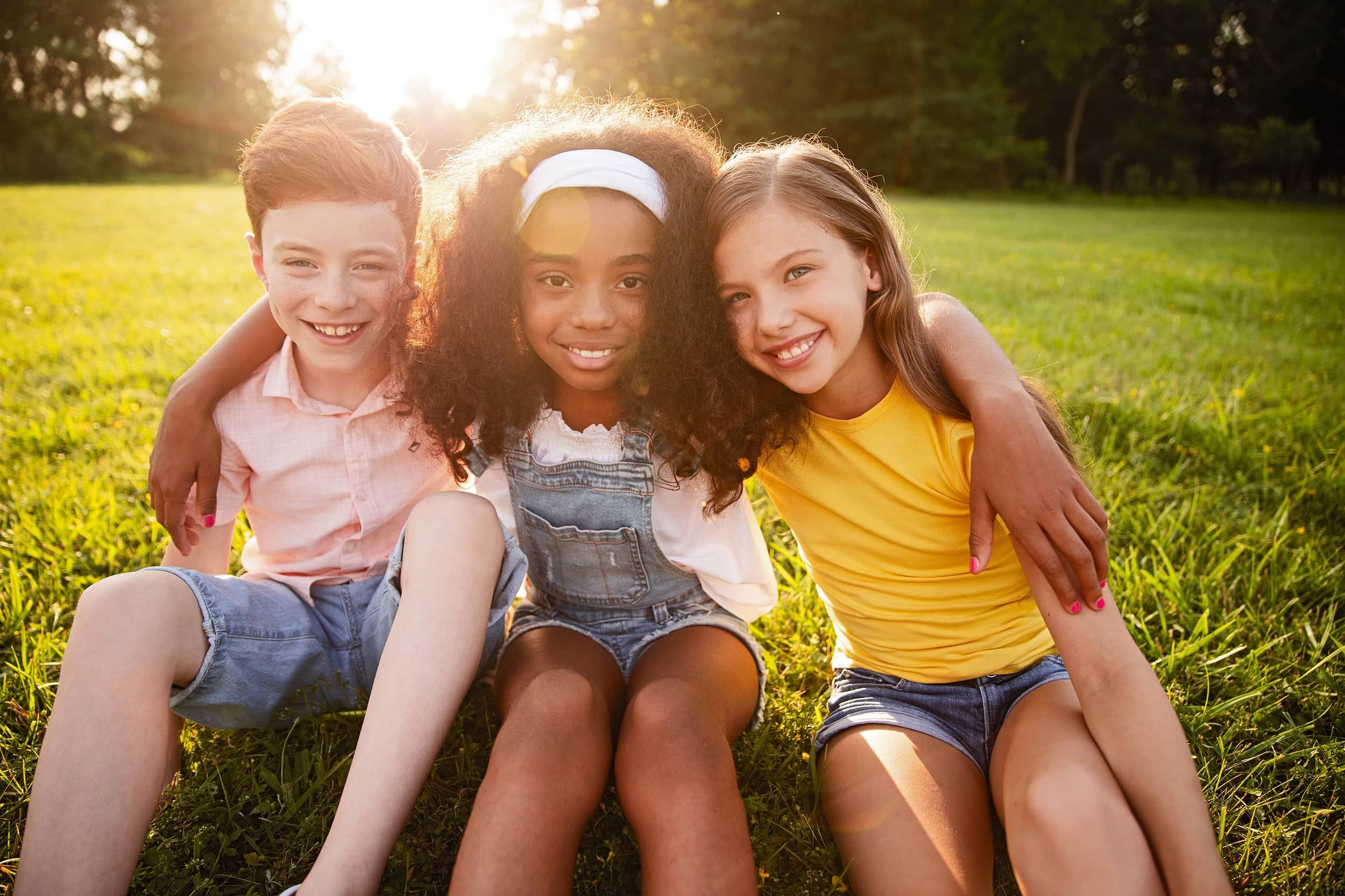 Three children sitting close together on a grassy field, smiling